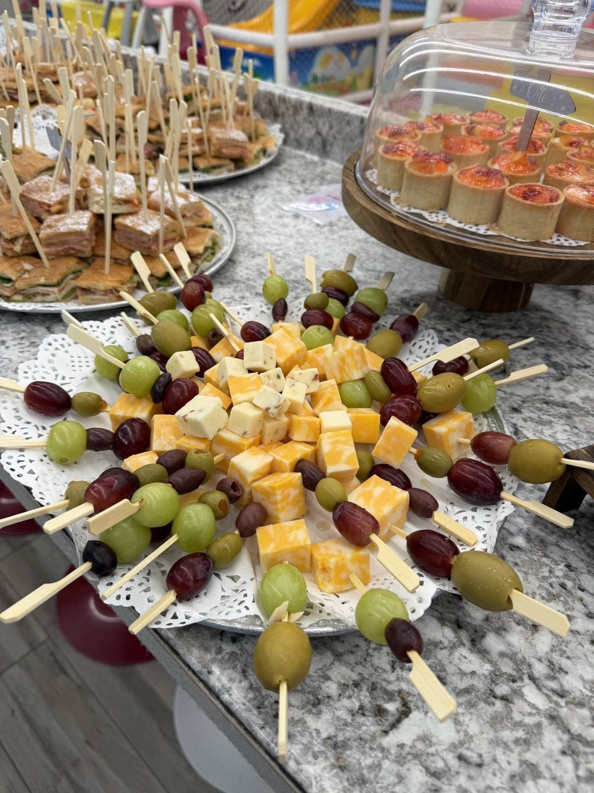 Catering spread at a private event — cheese, grape and olive skewers, club sandwiches, and mini tarts under a glass dome